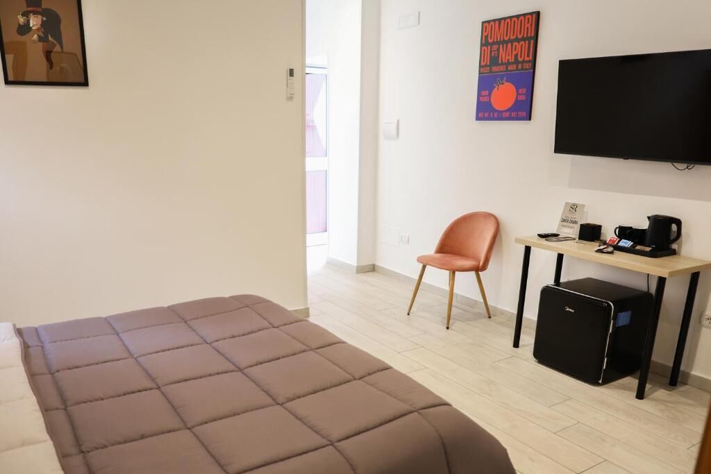 A room with a bed, a desk with a chair, a television, and a small black cabinet.