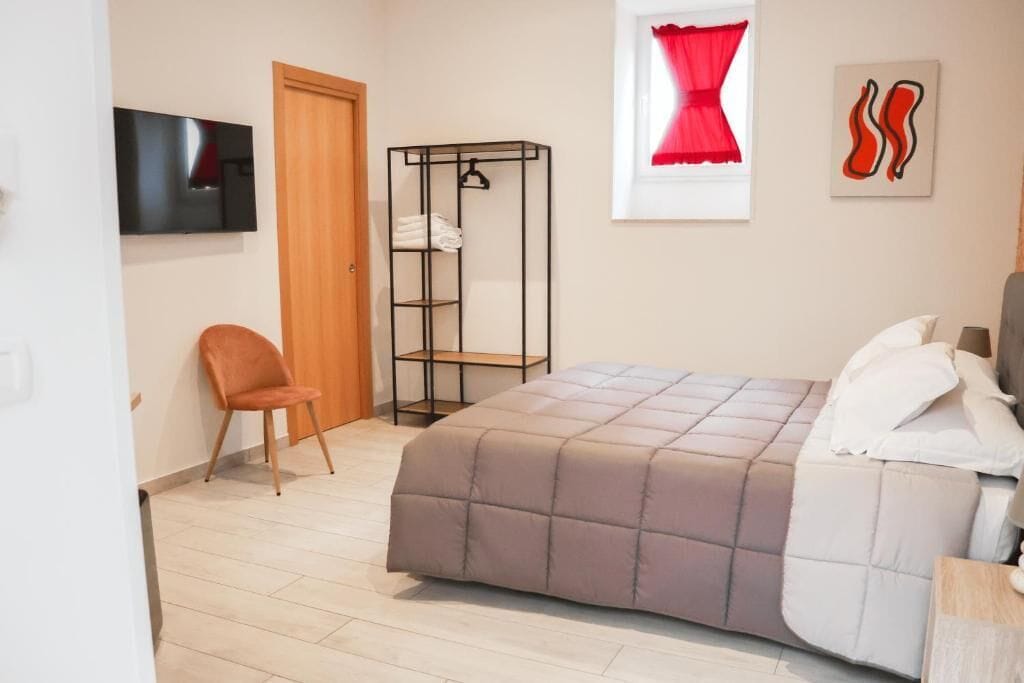 Une chambre à coucher avec un lit, une chaise, une armoire, un téléviseur et une fenêtre avec des rideaux.