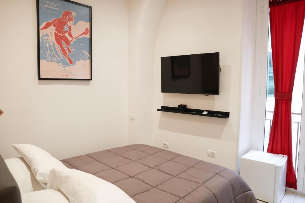 Une chambre à coucher avec un lit, un téléviseur fixé au mur, un rideau rouge et une affiche accrochée au mur.