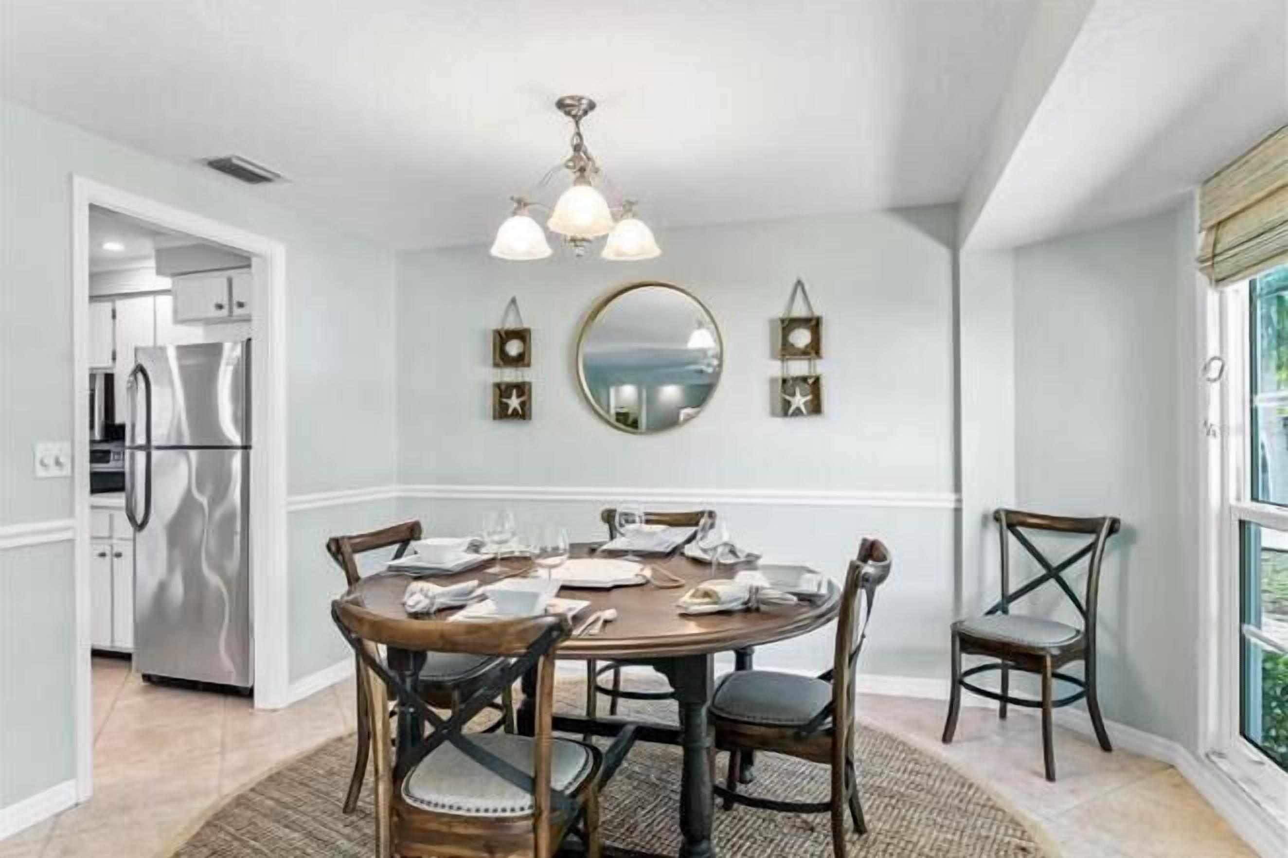 A dining area with a round table, chairs, a chandelier, a mirror, and a refrigerator in the background.