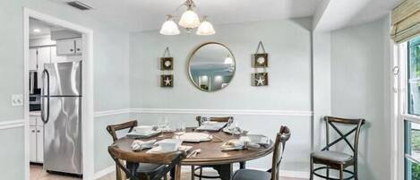 A dining area with a round table, chairs, a chandelier, a mirror, and a refrigerator in the background.