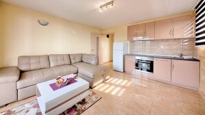 A modern living room with a beige sectional sofa, a white coffee table, and a kitchen area with a stove, oven, and cabinets. - Apartman ZIS 5L (Opština Bar)