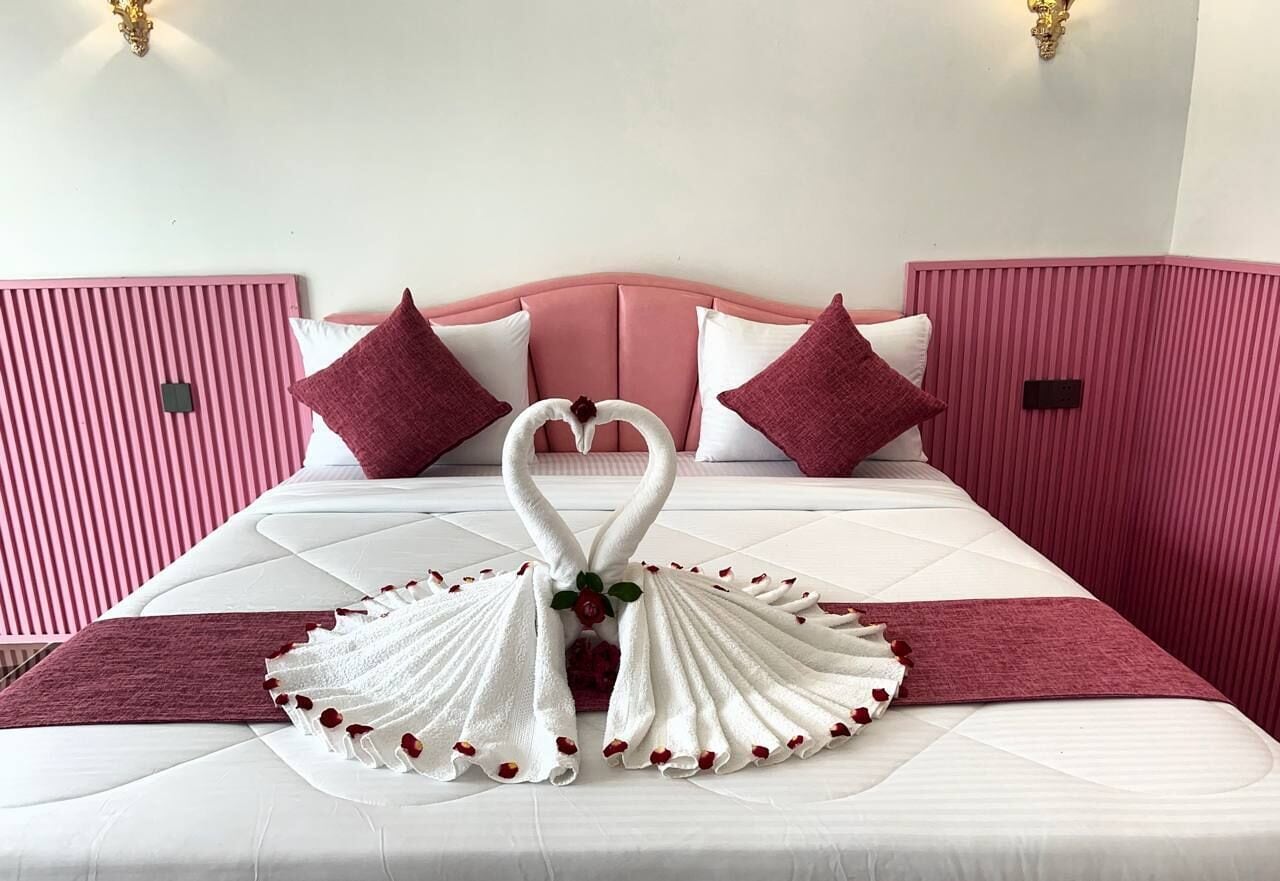 A bed with a pink headboard, white bedding, and a decorative towel arrangement in the shape of swans with roses.