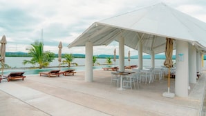 A covered outdoor seating area with lounge chairs and tables, overlooking a body of water.