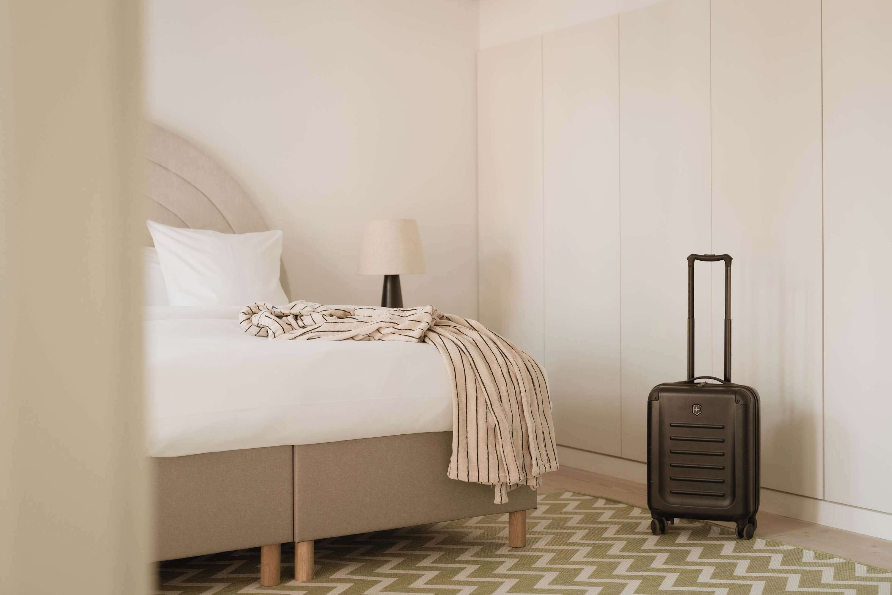Ein Bett mit einer gestreiften Decke, eine Lampe und ein Koffer in einem Hotelzimmer.