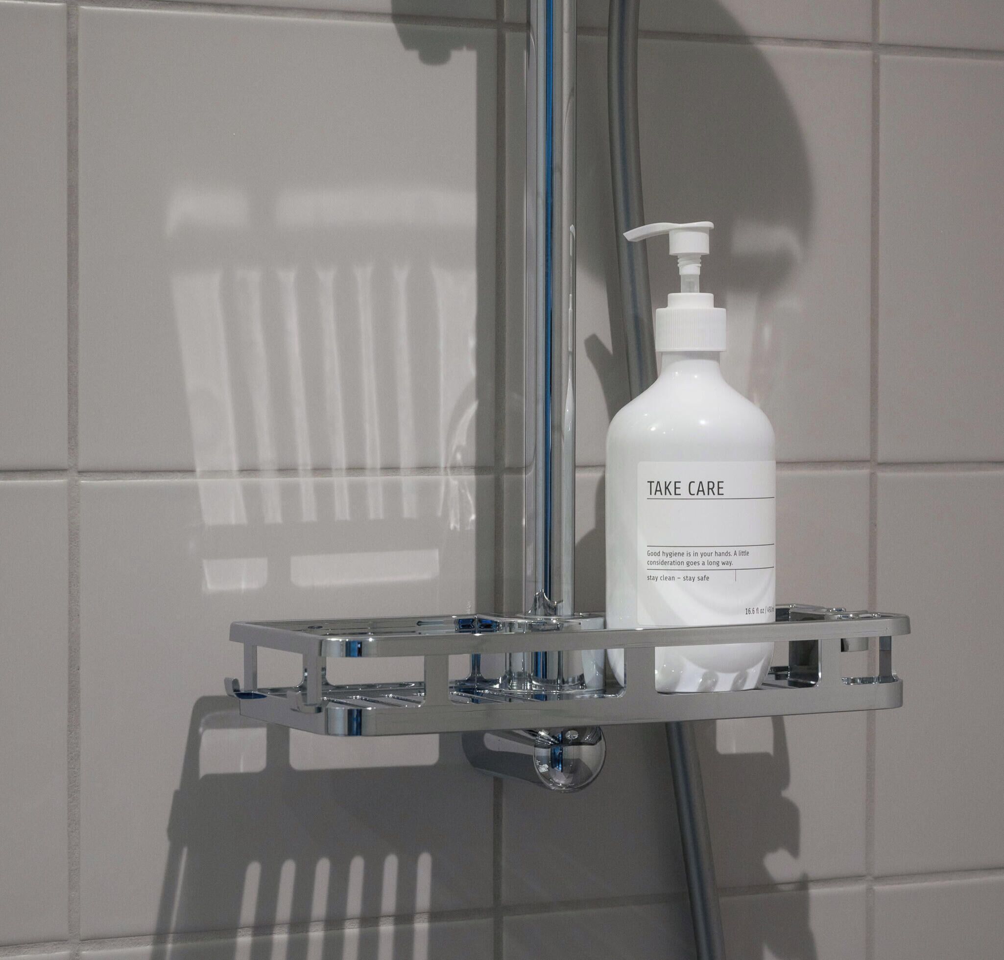 Een badkamer met een witte betegelde muur, een chroom douchemandje met een fles 'Take Care' handzeep, en een douchekop.