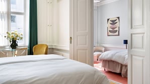 Una camera d'albergo con due letti, un angolo salotto con una poltrona, un tavolo con un vaso di fiori e vista sulla città attraverso una finestra con tende verdi.