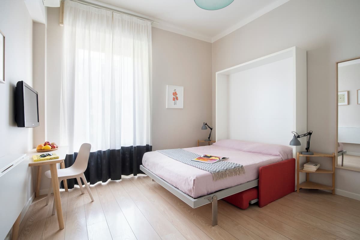 Quarto com cama, escrivaninha, cadeira, televisão e janela com cortinas.