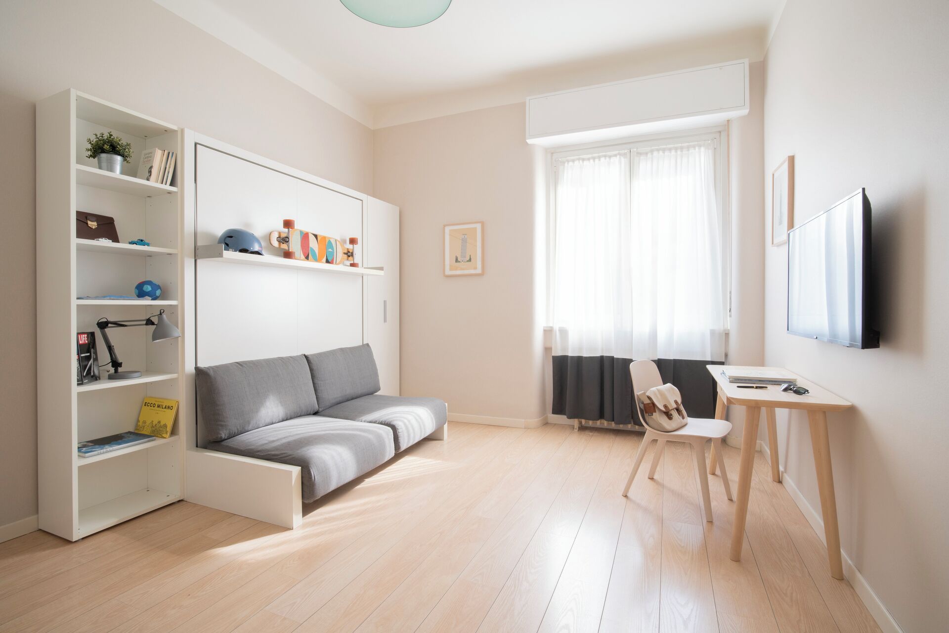 Ein modernes Wohnzimmer mit einem grauen Sofa, einer weißen Regaleinrichtung mit verschiedenen Gegenständen, einem kleinen Holztisch und einem Fenster mit transparenten Vorhängen.