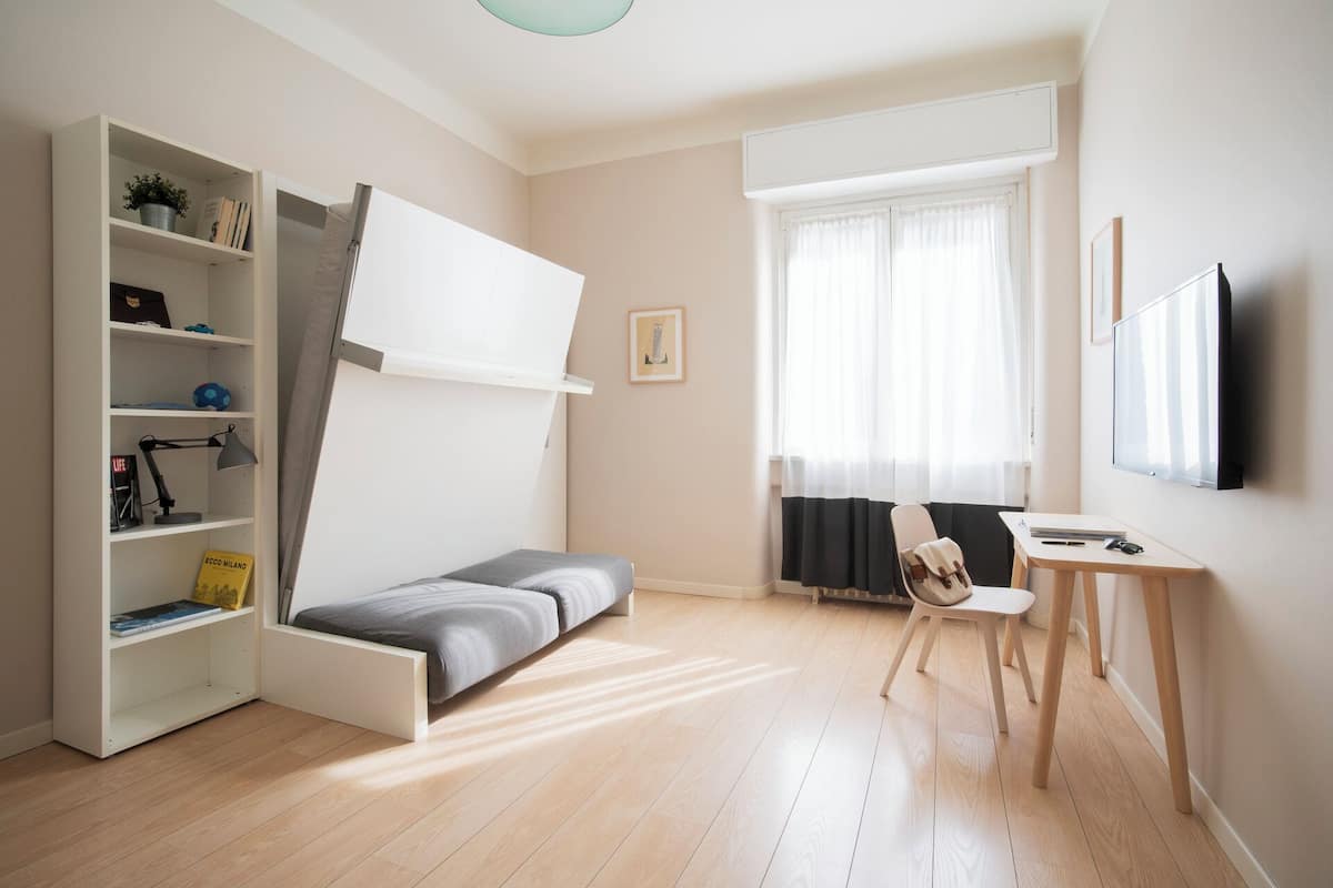 Quarto moderno com uma cama, uma escrivaninha, uma cadeira, uma estante de livros e uma televisão.