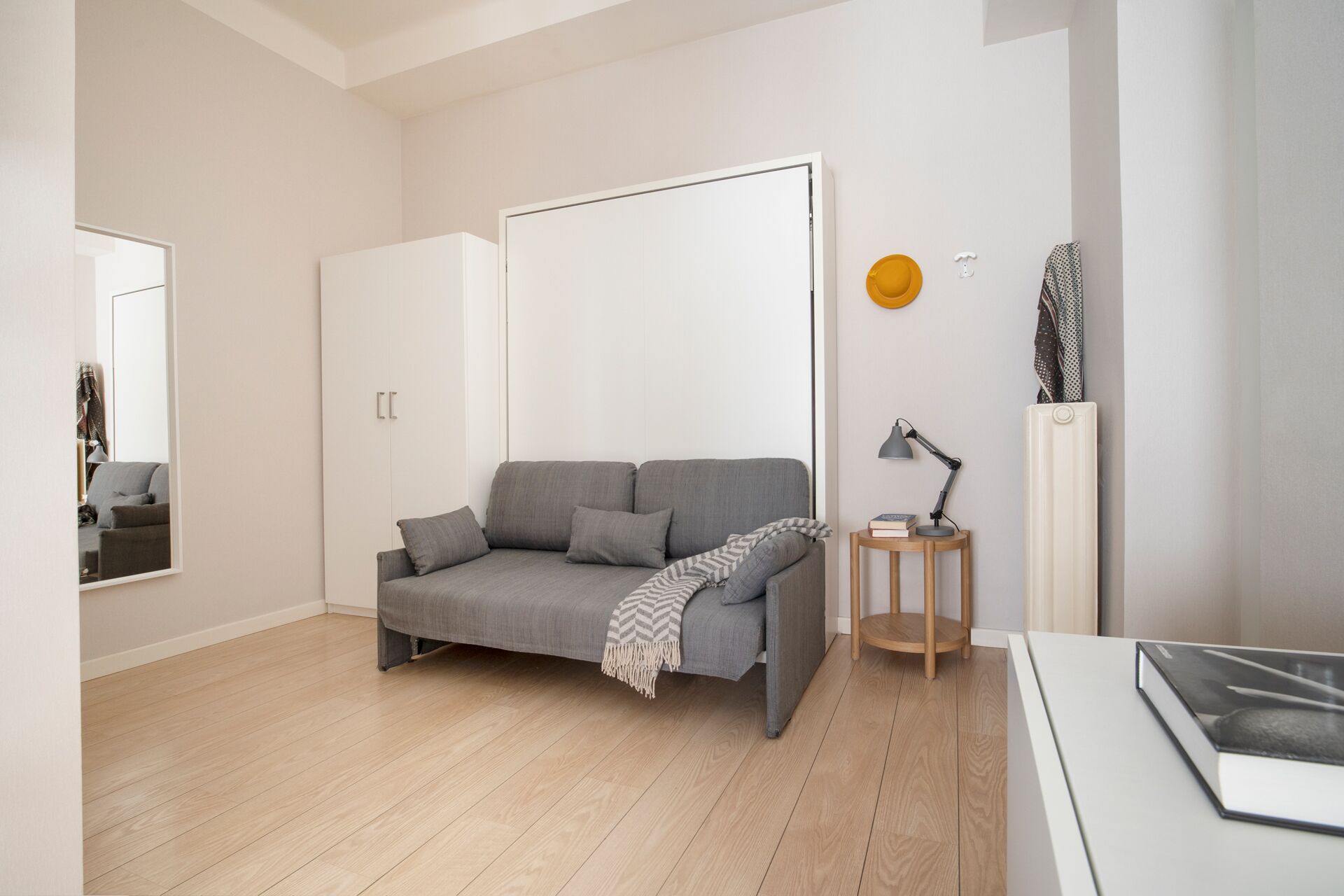 A modern living room with a grey sofa, a small wooden side table, a floor lamp, and a radiator.