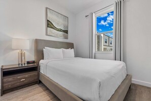 A hotel room with a bed, a nightstand, a lamp, a painting, and a window with a view of buildings. - Stylish 6BR townhome w/ private pool•Near parks (Davenport)