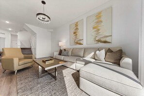 Una sala de estar moderna con un sofá, un sillón y una mesa de centro. Se observa una araña, una obra de arte en la pared y una escalera al fondo.