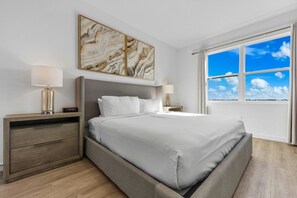 Un dormitorio moderno con una cama grande, dos mesitas de noche con lámparas, un pequeño mueble auxiliar y un ventanal con vistas al cielo.
