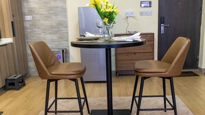 A dining area with two brown chairs, a table with a vase of yellow flowers, a glass of water, and a small refrigerator in the background.