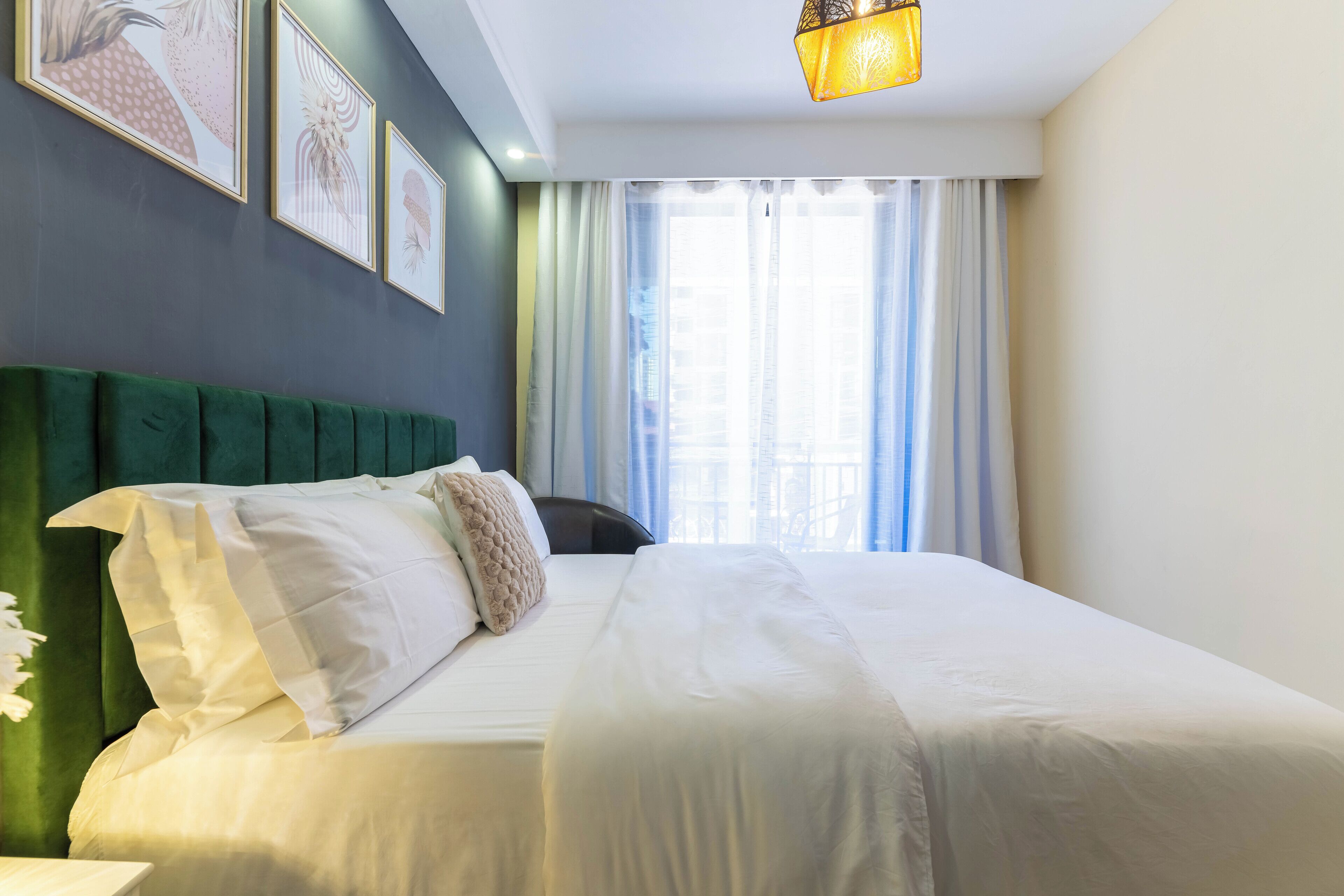 Un lit bien fait, avec du linge de lit blanc, une tête de lit recouverte d’un tissu vert et une fenêtre ornée de rideaux légers laissant filtrer la lumière naturelle.