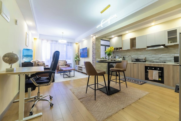A modern kitchen with a dining area, wooden flooring, and a large window providing natural light.