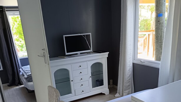 A compact room with a bed, a white desk, a wicker chair, a white cabinet with a TV, and a window with curtains.