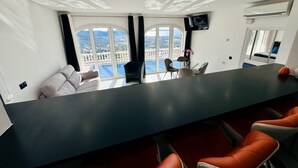 A modern hotel room with a large panoramic window, a dark bar area, and a seating arrangement with orange chairs. - Villa Serenity | Private pool, air conditioning, view of the bay of Roses, up to 13 people (El Mas Fumats)