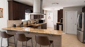 A modern kitchen with dark wood cabinets, a granite countertop, a stainless steel range hood, and a built-in oven. There are three bar stools and a large island with a sink.