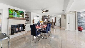 A spacious living room with a fireplace, a dining area with chairs, a kitchen in the background, and a ceiling fan.