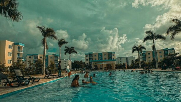 Una piscina con gente nadando y descansando, rodeada de palmeras y edificios de varios pisos.