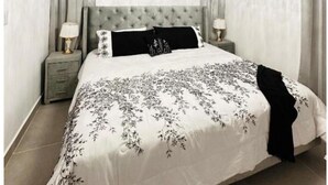 Un dormitorio con cabecera capitonada, una araña, una cama con colcha estampada y dos lámparas de noche en una mesita de noche gris.