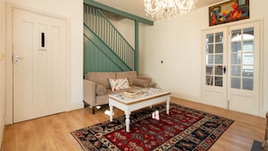 Un salon avec un canapé beige, une table basse blanche, un tapis à motifs, un lustre, un escalier avec des rampes de couleur turquoise et une porte avec des panneaux de verre.