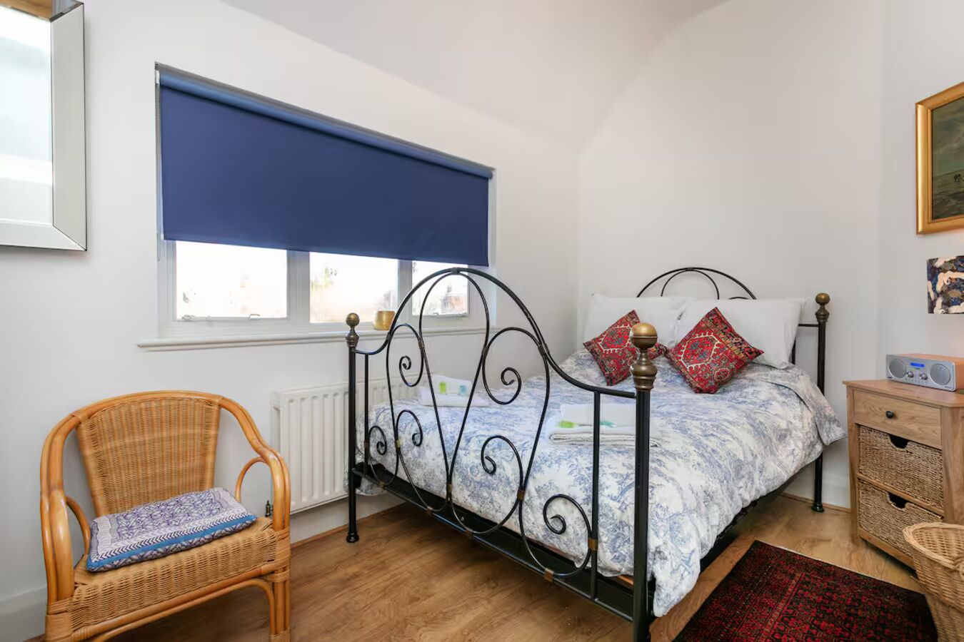 A bedroom with a metal bed frame, a wicker chair, a wooden nightstand, and a window with a blue blind.