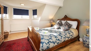 Une chambre avec un lit en bois, un tapis rouge à motifs, une commode en bois, un panier en osier et une petite table avec une lampe.