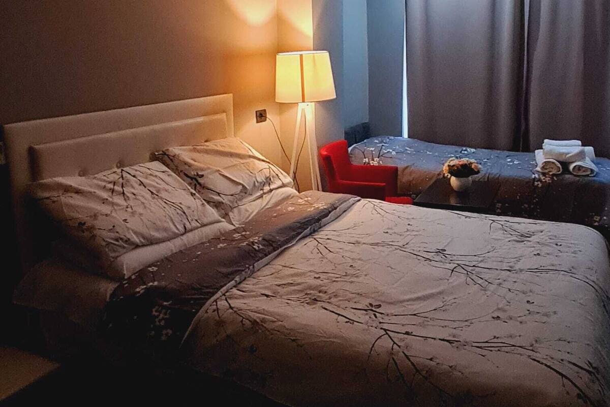 Une chambre d’hôtel avec un grand lit, un plus petit lit, un fauteuil rouge, une lampe et une fenêtre avec des rideaux.