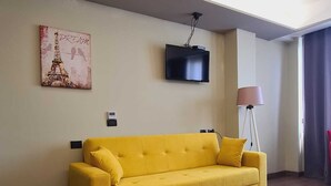 Un canapé jaune dans une pièce où l’on aperçoit un téléviseur à écran plat fixé au mur, une lampe et un tableau représentant la Tour Eiffel.