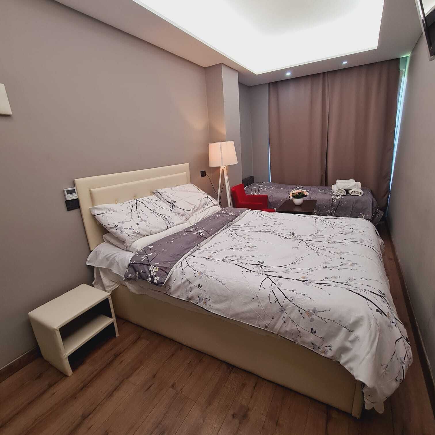 Un dormitorio con una cama grande, una mesita de noche, una silla roja, un escritorio con una lámpara y vistas al cielo a través de un tragaluz.