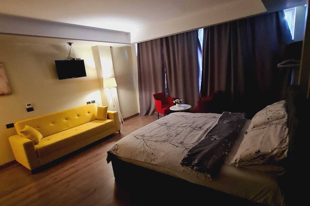Une chambre d’hôtel avec un lit, un canapé jaune, une petite table avec un vase et un téléviseur fixé au mur.