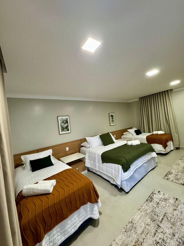 A hotel room with three beds, each with a different colored bedspread, a wooden headboard, and a small bedside table with a lamp.