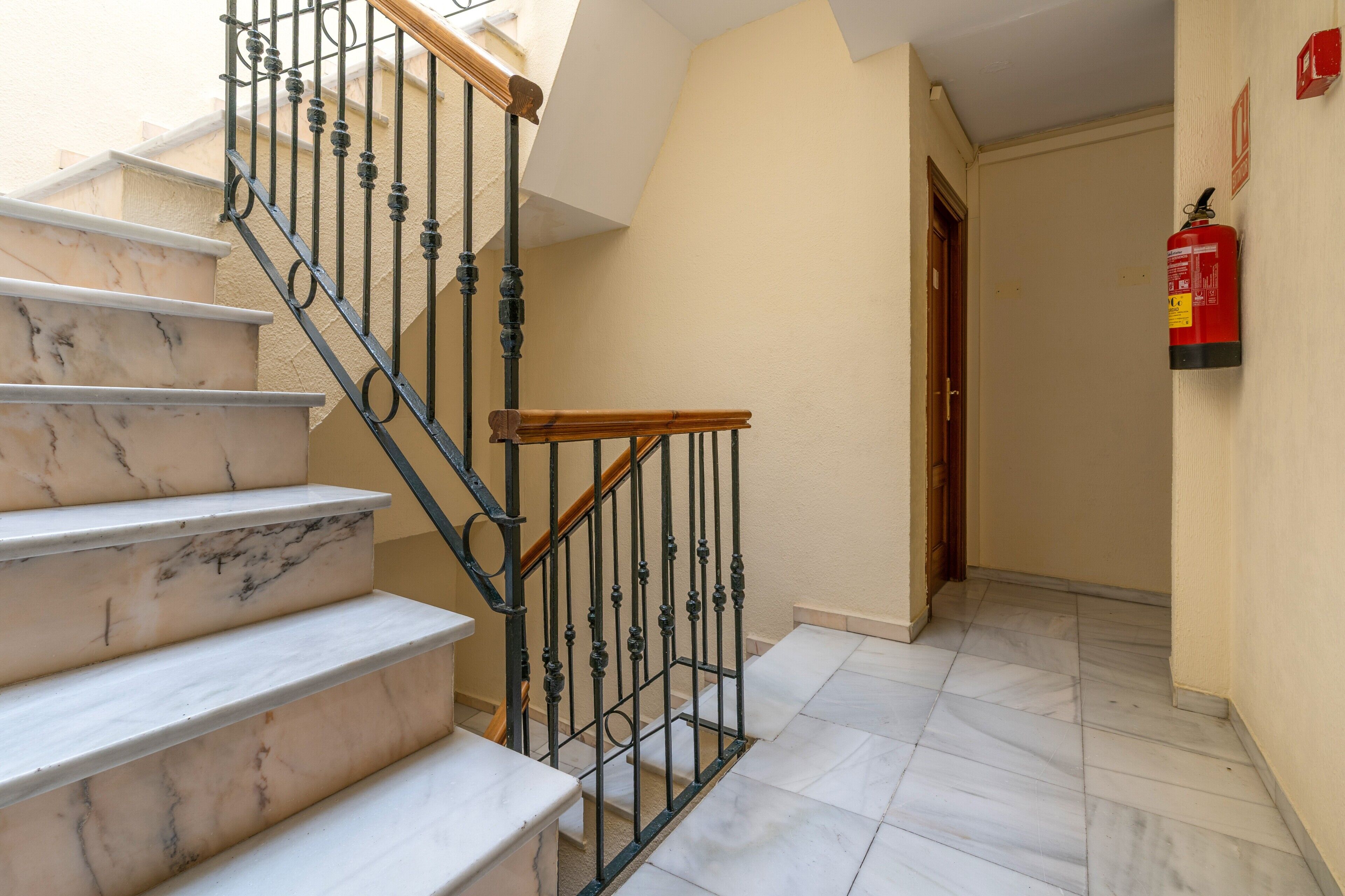Un escalier avec des marches en marbre et des rampes en métal noir, menant à une pièce dotée d’une porte et d’un extincteur fixé au mur.