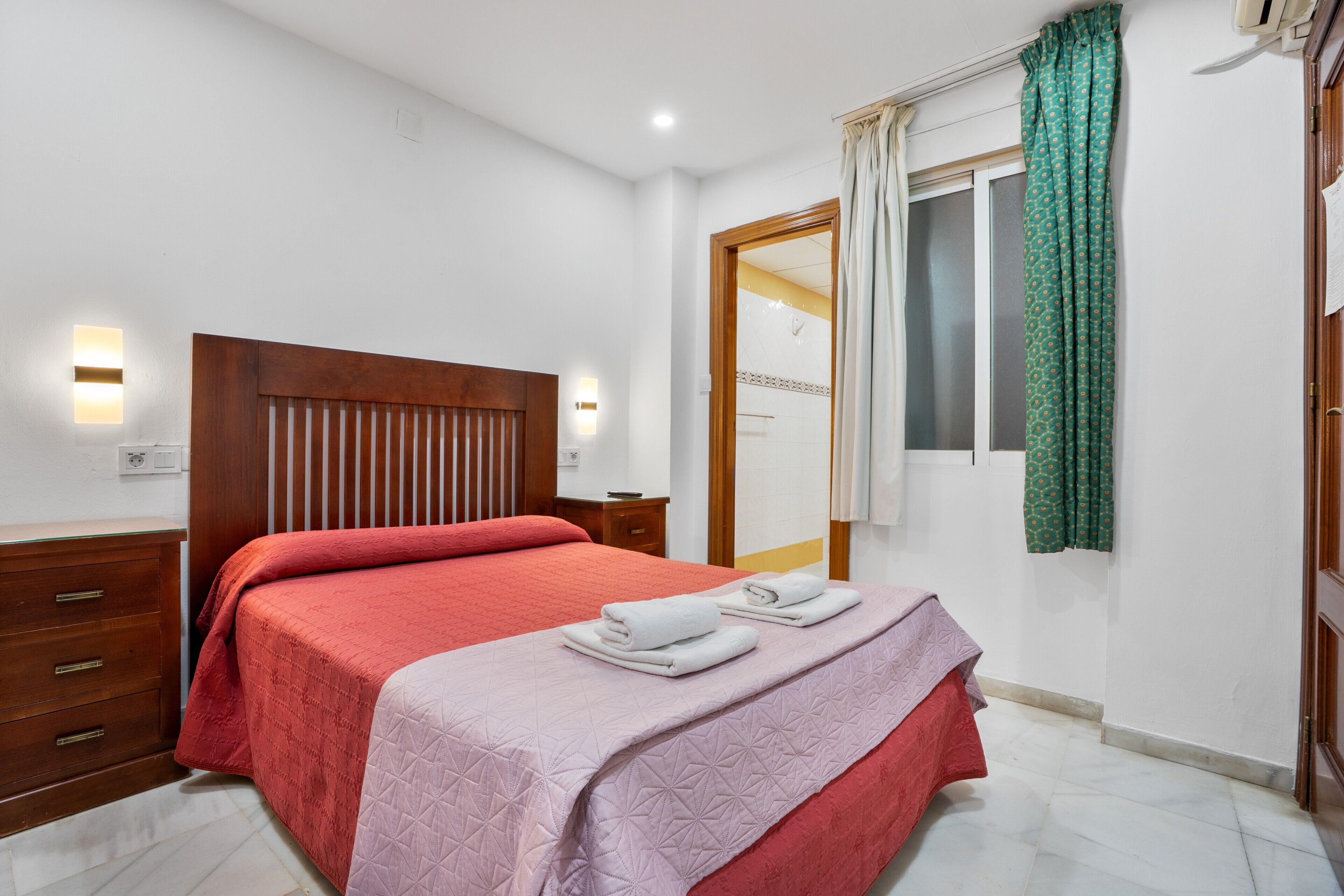 A bedroom with a wooden headboard, a bed with red and pink bedding, two towels on the bed, a wooden nightstand, and a mirror with a green curtain.