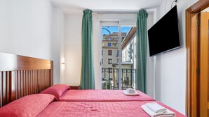 Ett hotellrum med en säng, en tv, ett fönster med gardiner och utsikt över byggnader.