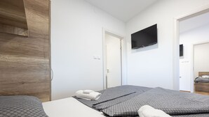 A modern hotel room with a wooden wardrobe, a flat-screen TV mounted on the wall, and a neatly made bed with grey bedding and white towels. - Camping Karin - Mobile houses Magoro (Donji Karin)