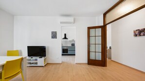 Une pièce avec un plancher en bois, un téléviseur posé sur un meuble, une chaise jaune et une table blanche.