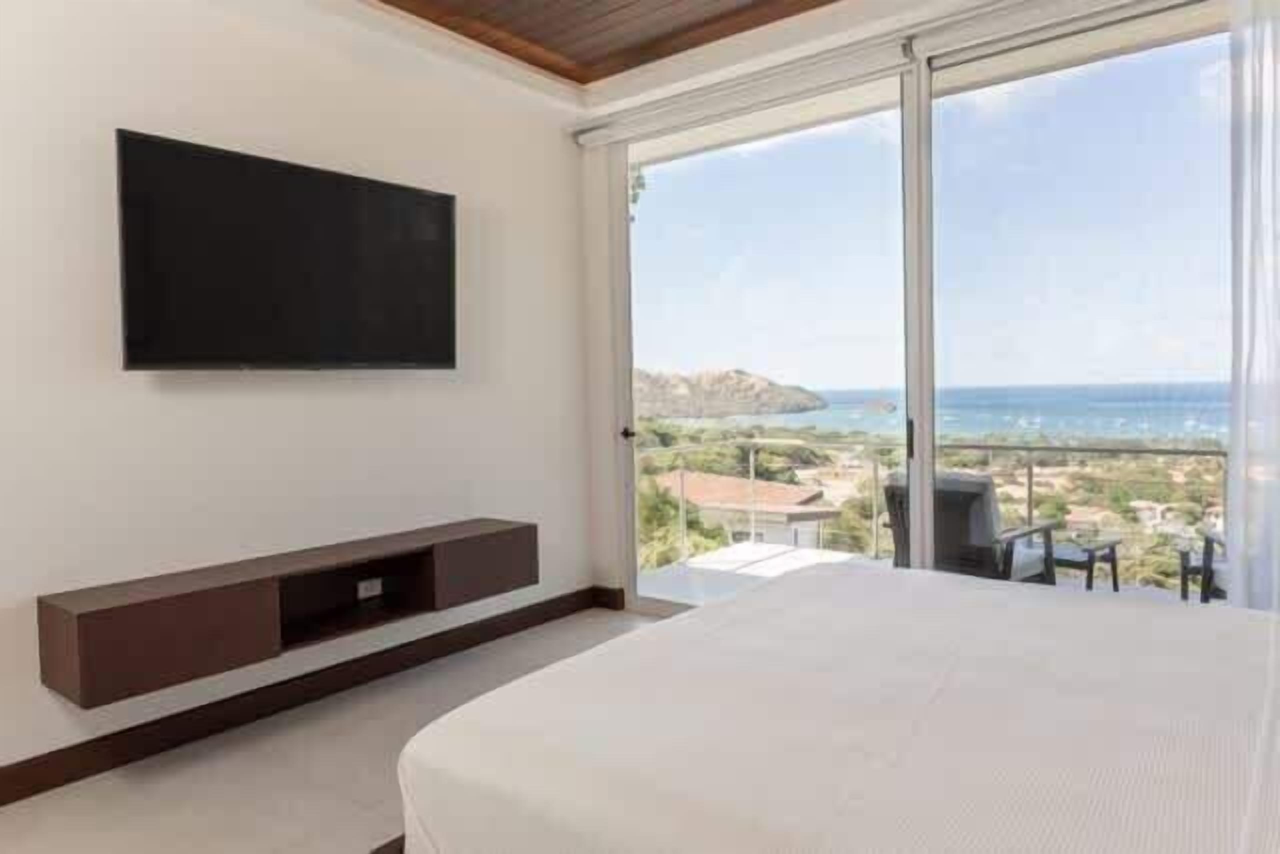 Ein Schlafzimmer mit einem großen Bett, einem an der Wand befestigten Fernseher und einer Schiebetür, die auf einen Balkon mit Blick auf das Meer und die Stadt führt.