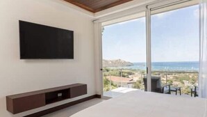 Un dormitorio con una cama grande, un televisor montado en la pared y un ventanal corredero que da a un balcón con vistas al mar y edificios.