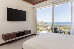 A bedroom with a large bed, a TV mounted on the wall, and a sliding glass door leading to a balcony with a view of the sea and buildings.