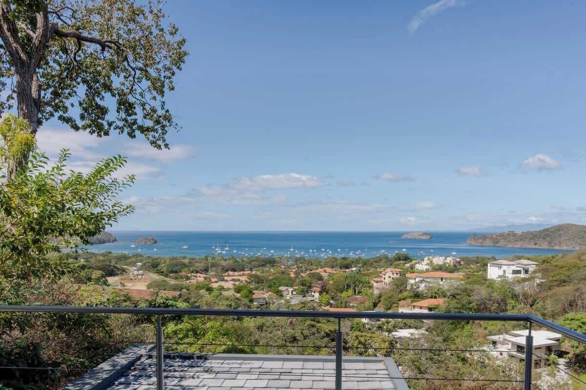 A balcony with a view of a coastal town, a tree, and a clear sky.
