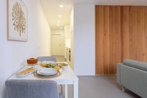 Ein zeitgemäßer Wohnbereich mit Essbereich, inklusive Tisch, gedeckt mit Brot, Obst und einem Glas Orangensaft. Ein graues Sofa und eine Holzpaneelwand sind zu sehen.