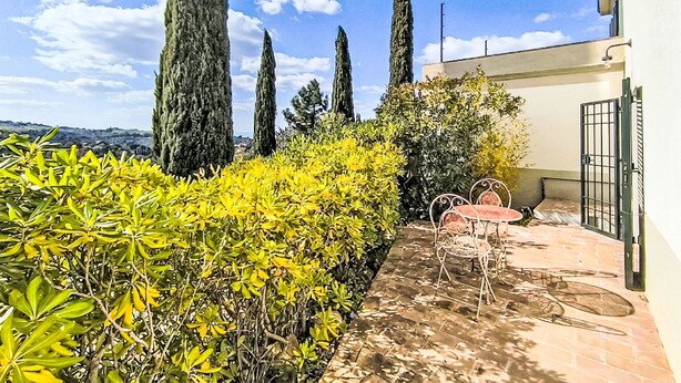 Un patio con mesa y sillas, rodeado de follaje y árboles altos.