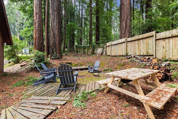 Ein rustikaler Außenbereich mit Adirondack-Stühlen und einem Picknicktisch, umgeben von einer Holzterrasse, inmitten eines bewaldeten Gartens mit hohen Bäumen und einem Holzzaun.