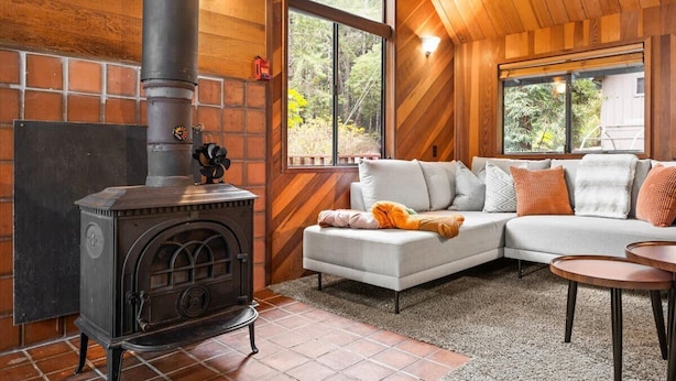 A cozy living room with a wood-burning stove, a sectional sofa, and large windows offering a view of the outdoors.