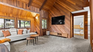 A cozy living room with a wooden ceiling, a sofa with cushions, a coffee table, and a TV mounted on the wall.