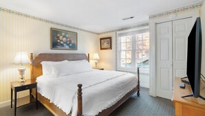 A neatly arranged bedroom with a wooden bed, two bedside tables with lamps, a television on a wooden dresser, and a window with a view of the outside. - Cottage 9 – Downtown Jacksonville Retreat Steps from Shops & Britt Gardens (Jacksonville)